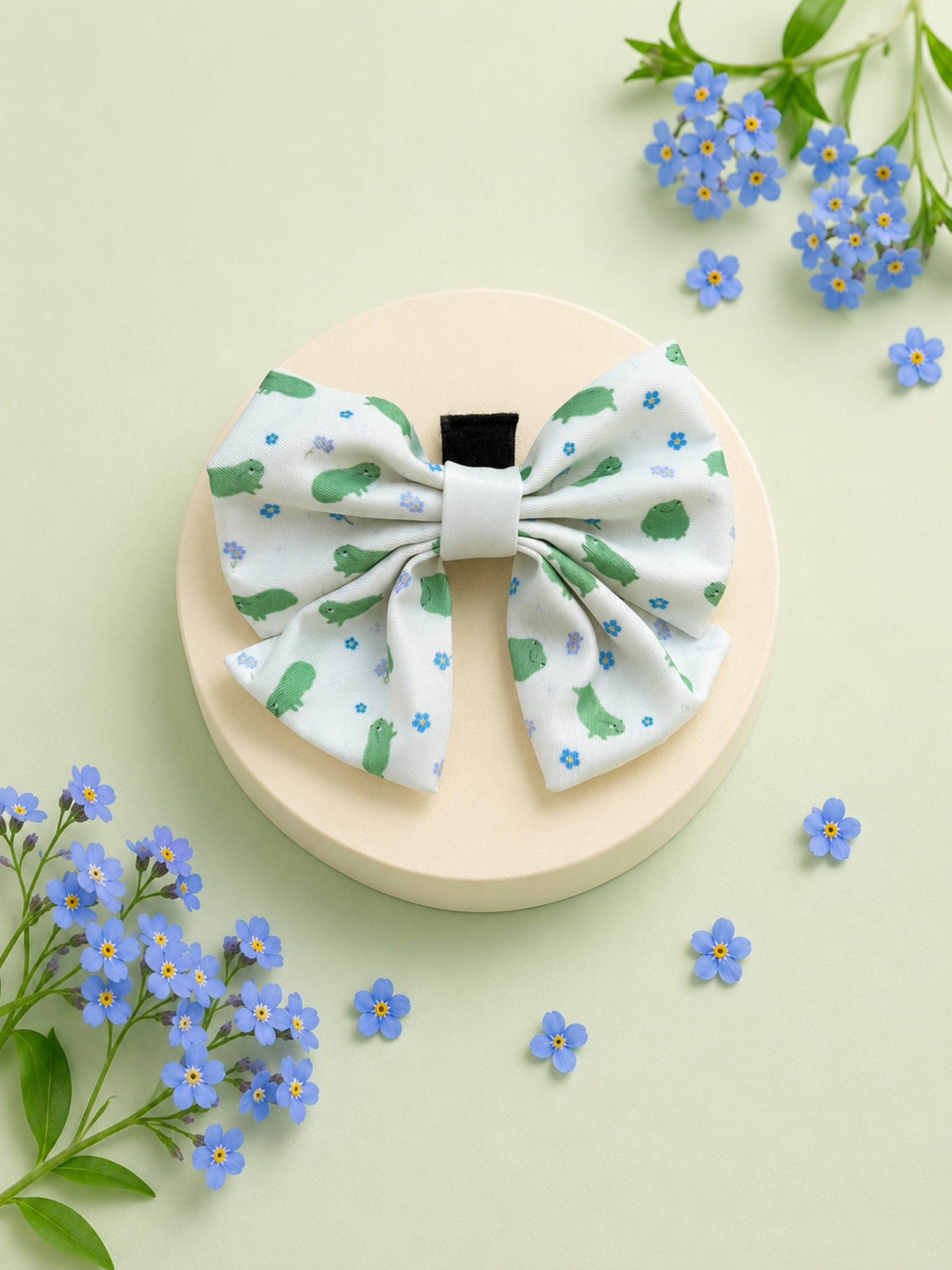 A fabric dog bowtie with a green frog and blue flower print on a white background, displayed on a round beige pedestal with decorative forget-me-not flowers.