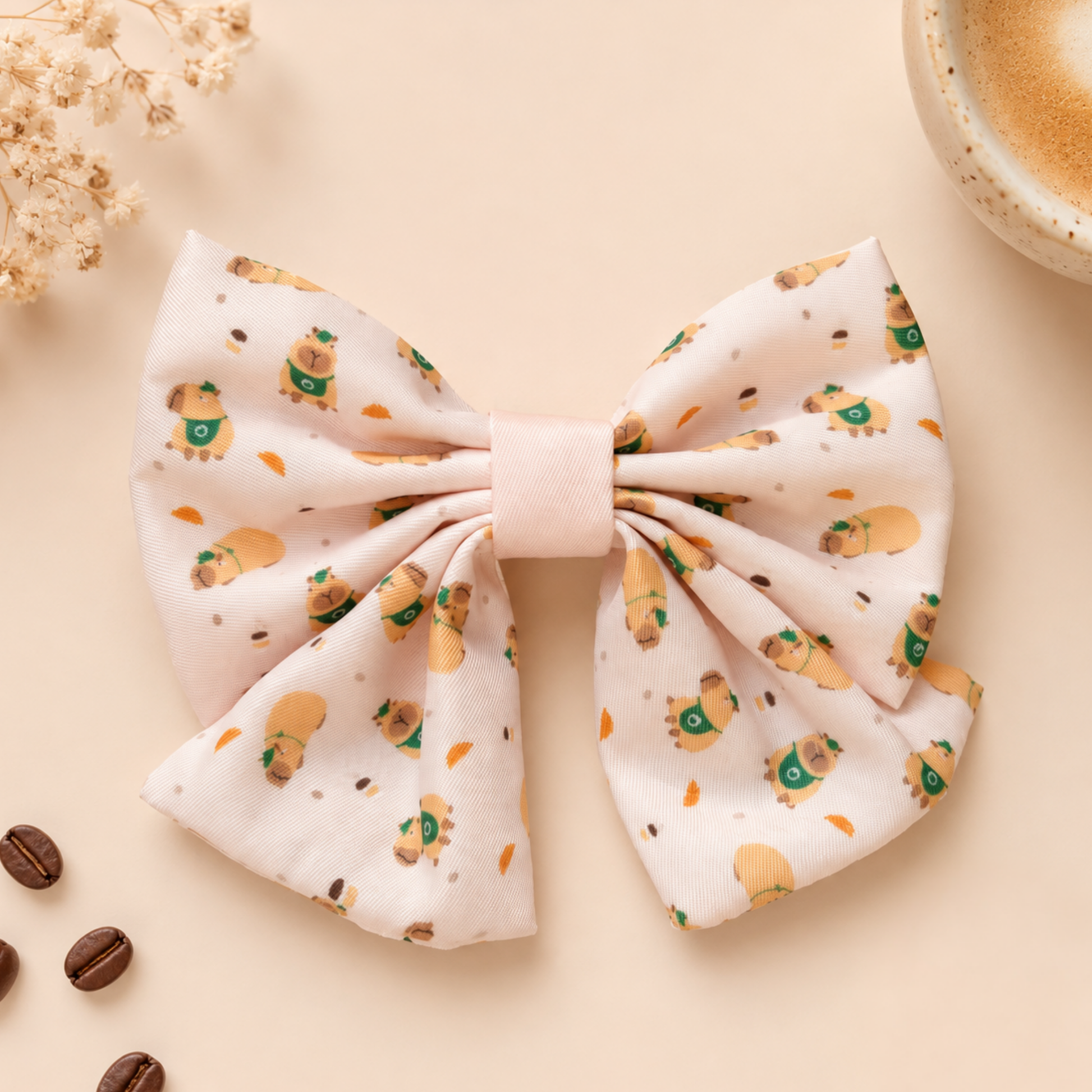 A velcro dog bow with a light pink base and a pattern of small cartoon capybara in green and orange, styled on a beige background with coffee beans and a cup of coffee.