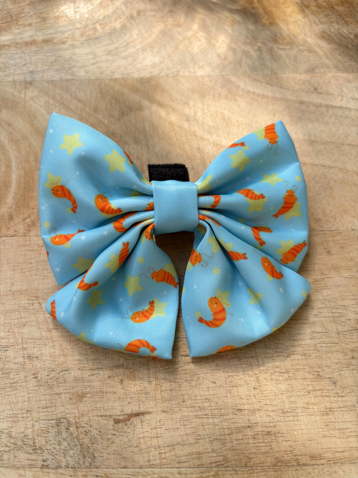 A light blue dog bowtie with an orange shrimp and yellow star pattern, displayed on a wooden surface.
