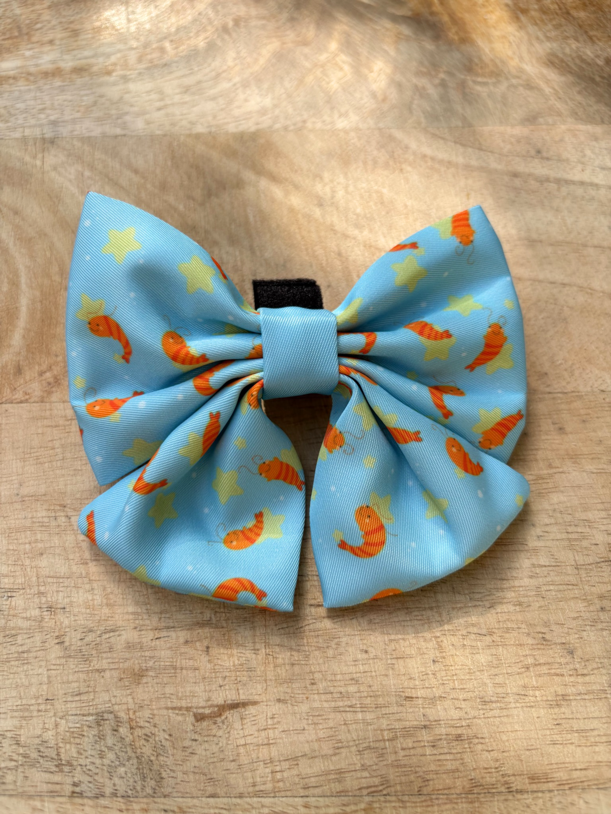 A light blue dog bowtie with an orange shrimp and yellow star pattern, displayed on a wooden surface.