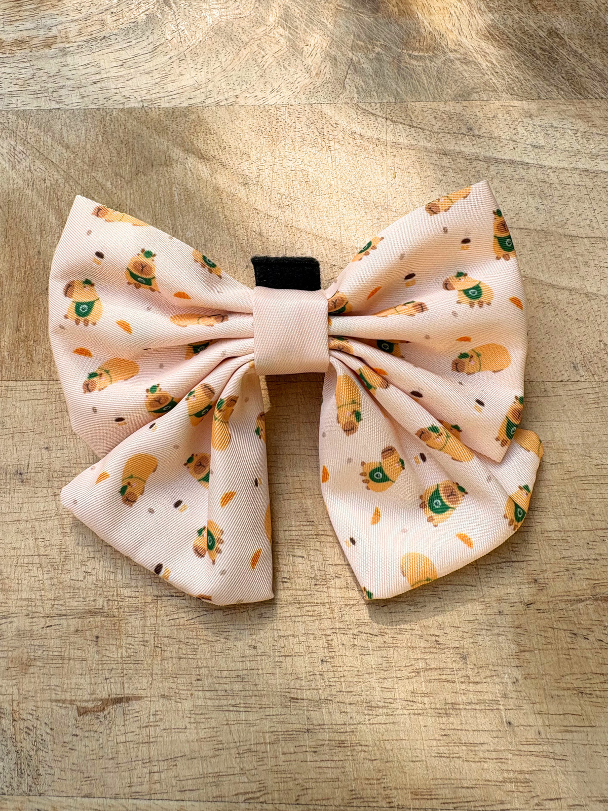 A light pink fabric bow for dogs with a pattern of small cartoon animals and leaves, displayed on a rustic wooden surface.
