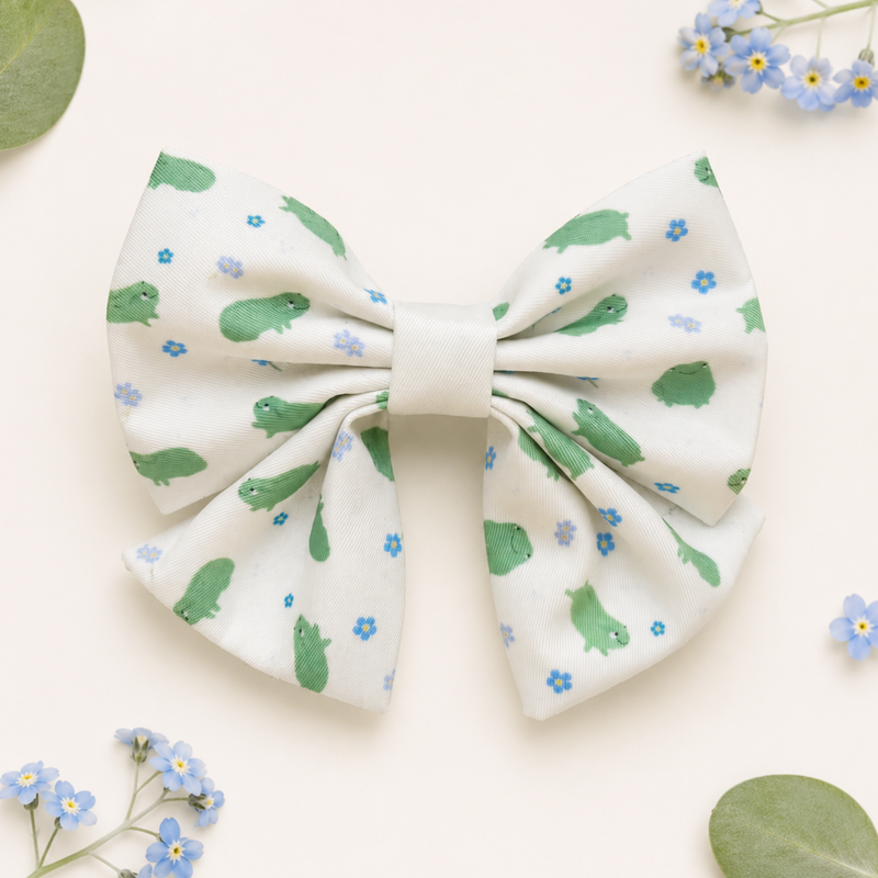 A dog collar bowtie featuring a white background with a pattern of green frogs and small blue flowers, styled on a light cream background with decorative green leaves and forget-me-not flowers.
