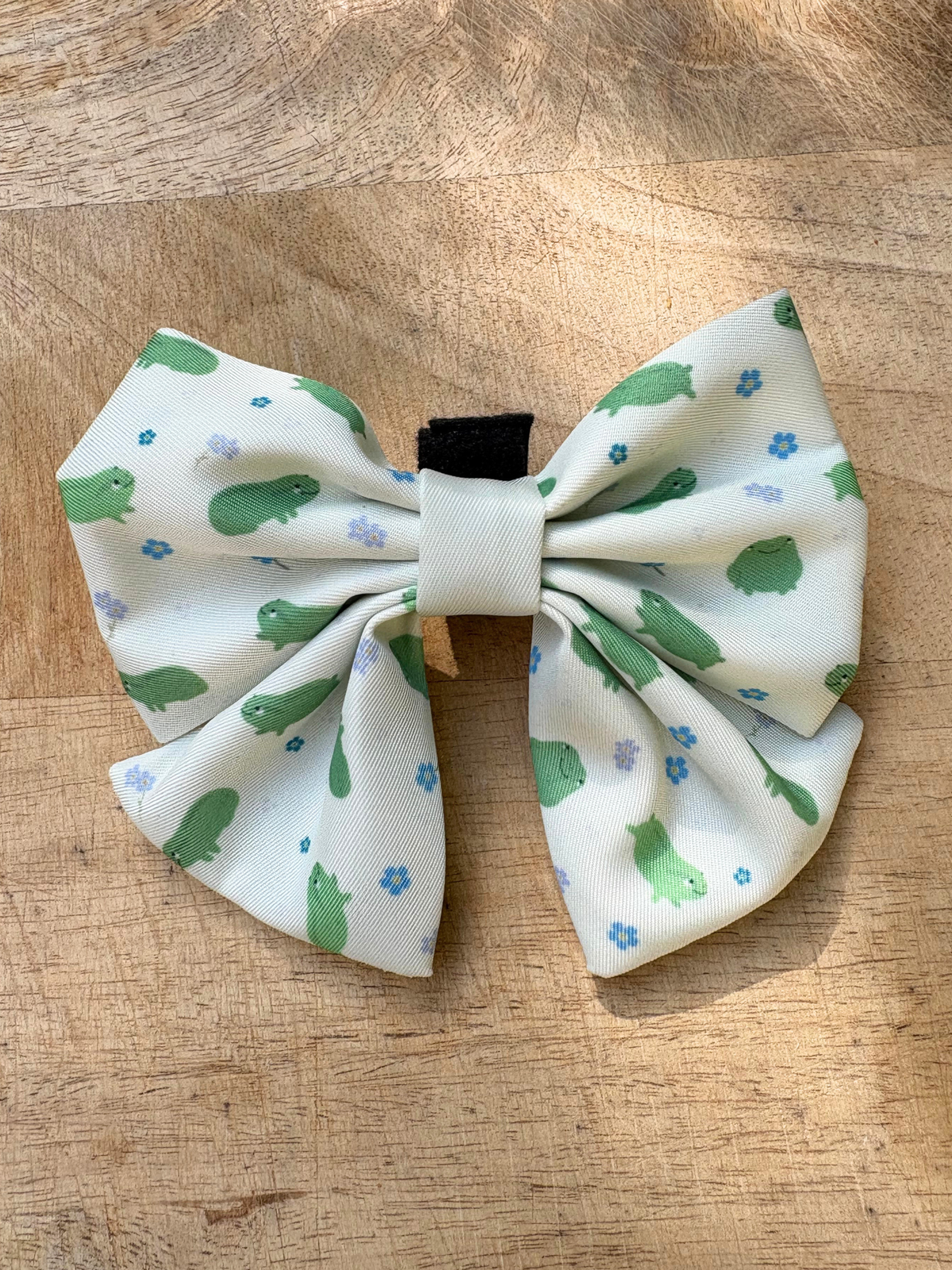 A fabric pet collar bow featuring a white background with a print of green frogs and small blue flowers, featuring a velcro strap and displayed on a wooden surface.