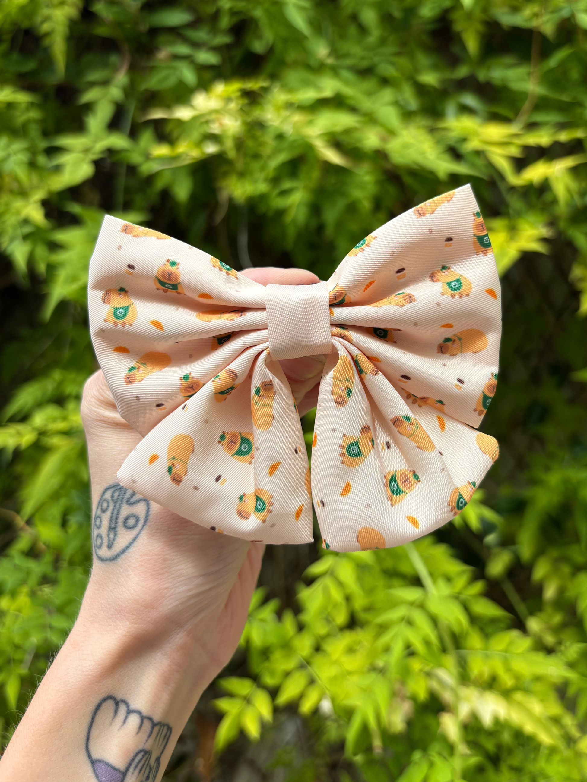 A hand holds a dog collar sailor bow featuring a light beige background with a playful orange-brown cartoon pattern and croissant print, set against a blurred background of green foliage.