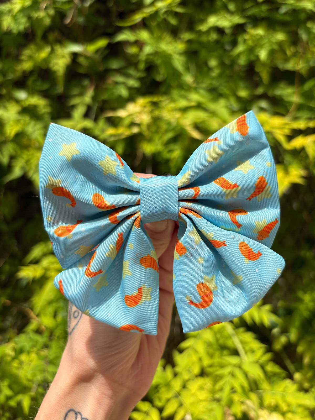 A light blue cute fabric dog sailor bow with an orange shrimp and yellow star pattern, held by a hand against a blurred green foliage background.