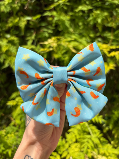 A light blue cute fabric dog sailor bow with an orange shrimp and yellow star pattern, held by a hand against a blurred green foliage background.