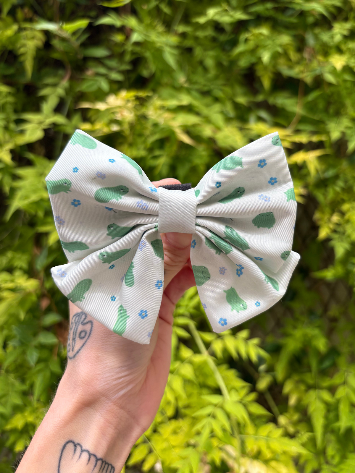 A hand holds a kawaii dog sailor bow featuring a white background with green frog and blue flower patterns, set against a blurred green foliage background.