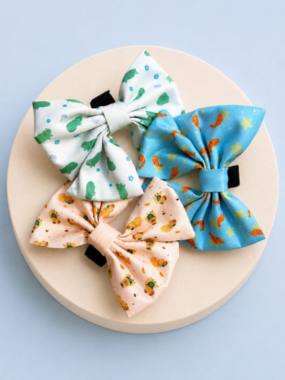 Three dog collar bows with different animal-themed prints are arranged on a round beige pedestal against a light blue background. The top-left bow is white with green frog and floral print, the bottom-left bow is peach with orange capybara and coffee print, and the top-right bow is light blue with orange shrimp and yellow stars patterns