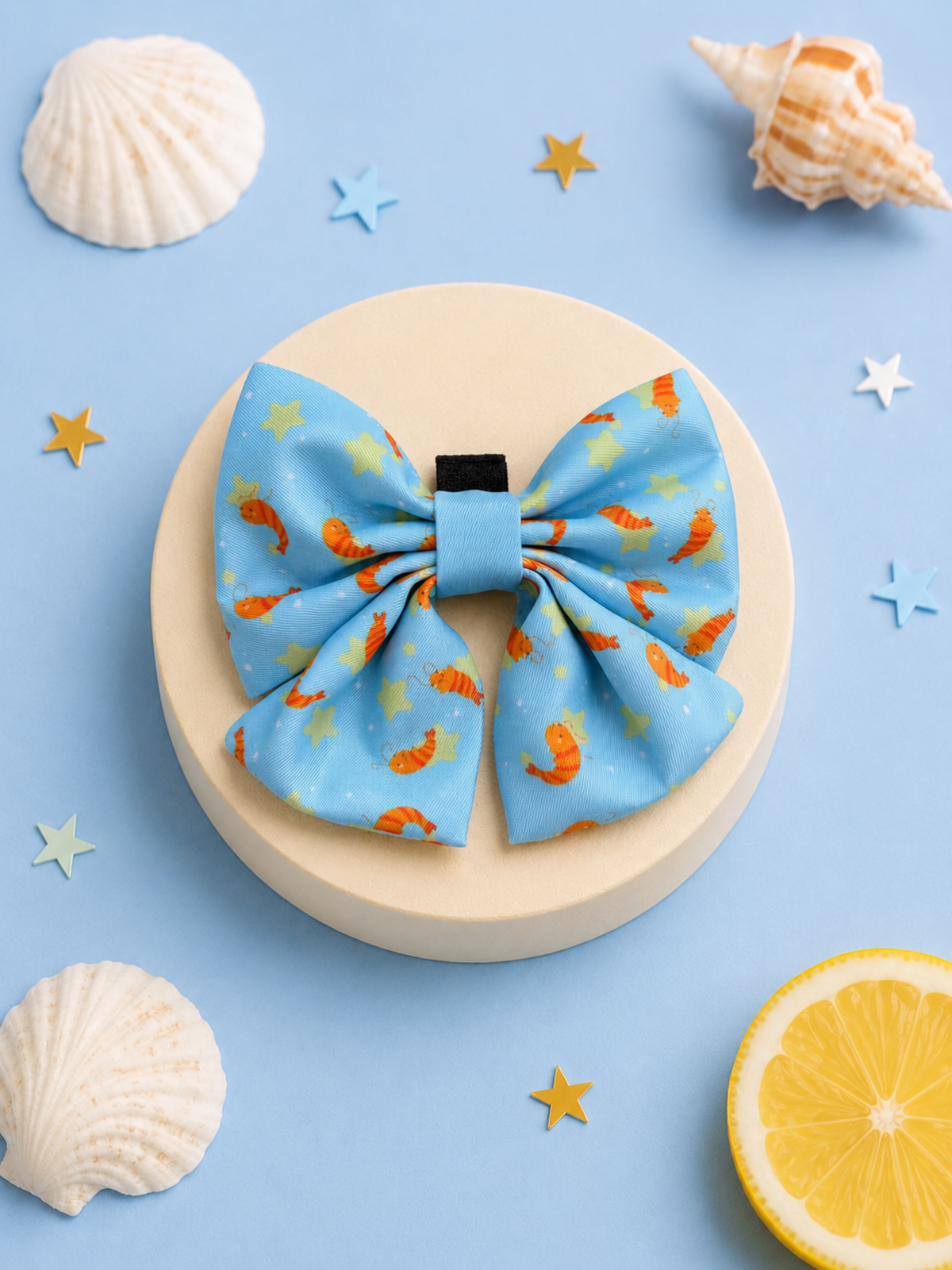 Blue Prawn Star sailor bow for dogs with an orange and yellow seashell pattern, displayed on a round beige pedestal with seashells and a lemon slice on a light blue background.